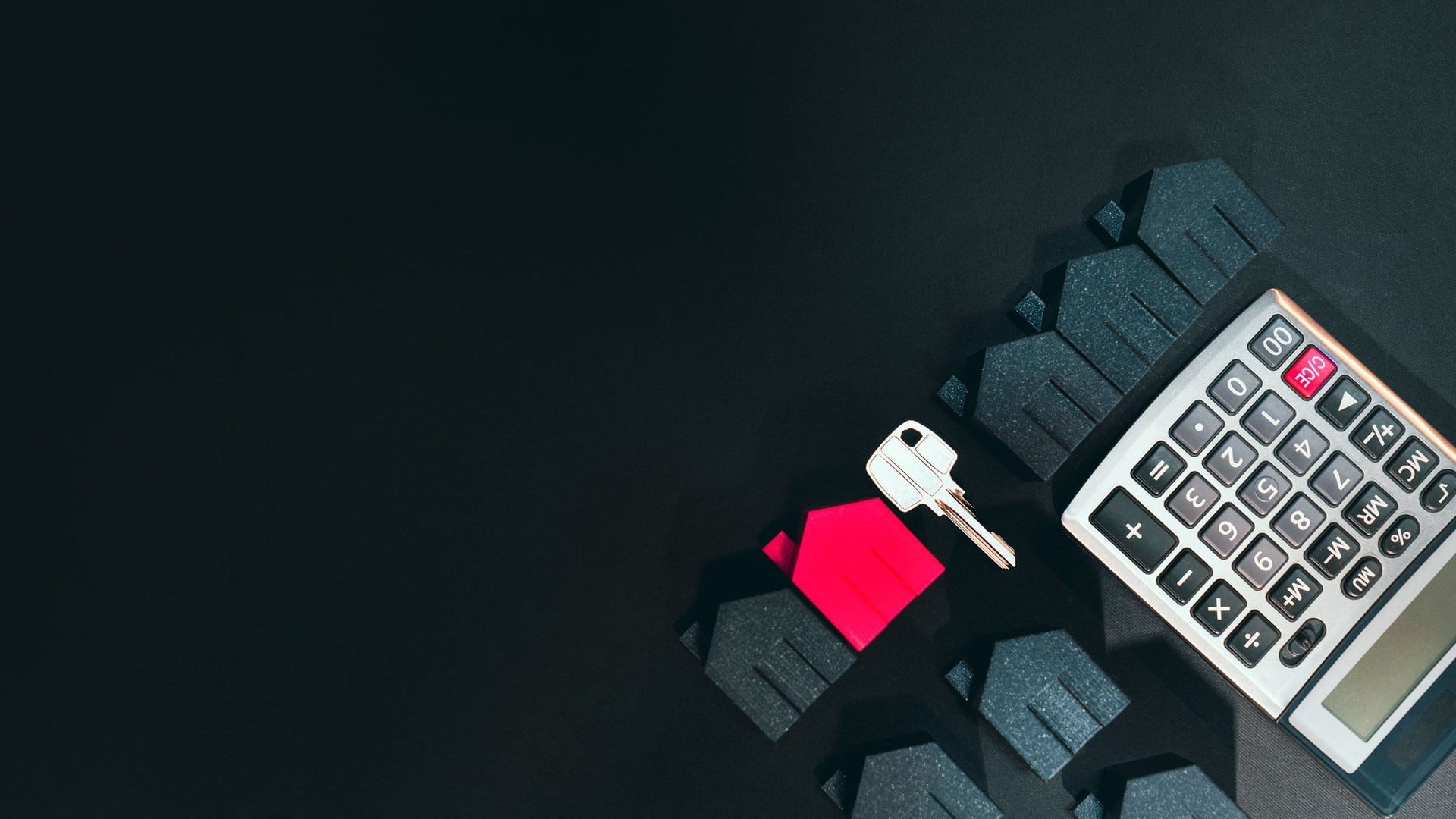 A calculator, a silver key and small dark model houses, one of them red, on a black surface.