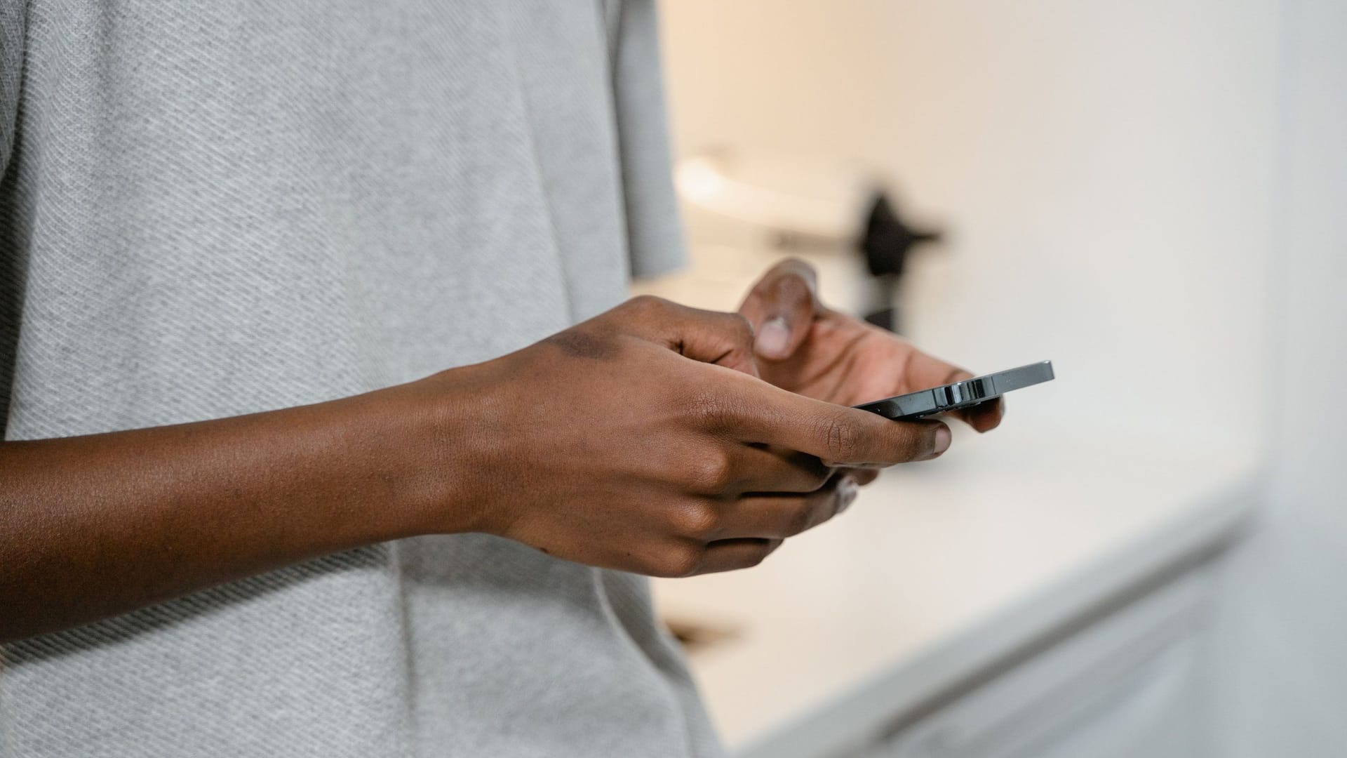 A person in a gray shirt types on a black smartphone, a kitchen counter blurred behind them.