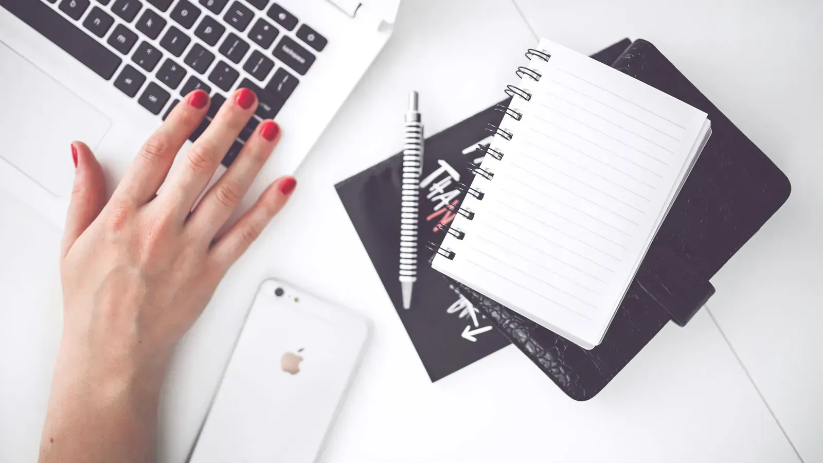 A hand with red nail polish rests on a white laptop keyboard beside a white phone, a striped pen and a blank spiral notebook.