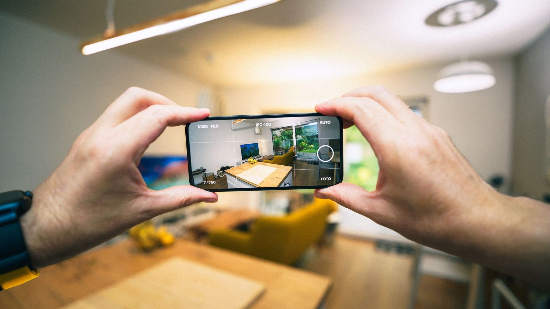 Two hands hold a smartphone sideways to photograph a bright open-plan room, the screen showing a kitchen island, a TV and a yellow sofa.