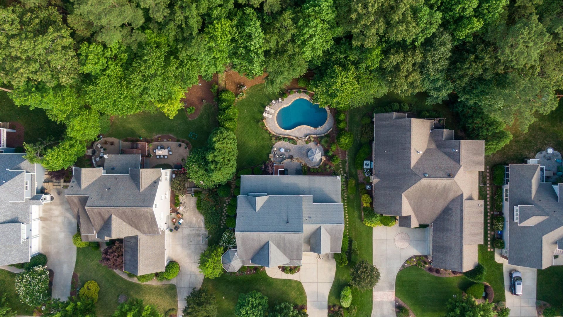 Aerial view of similar suburban houses with gray roofs, driveways and one backyard pool, surrounded by trees.