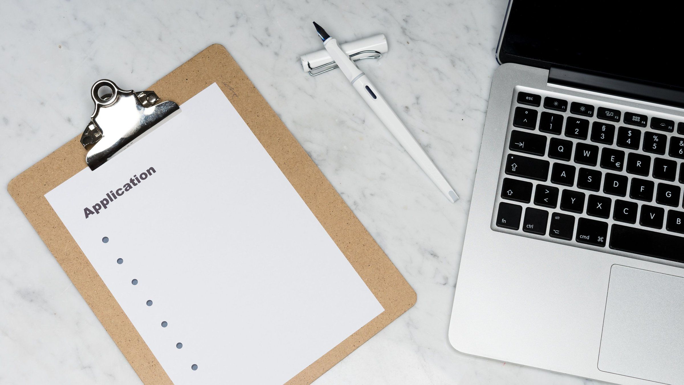 A blank application form on a wooden clipboard lies on a marble desk beside a white fountain pen and an open laptop.